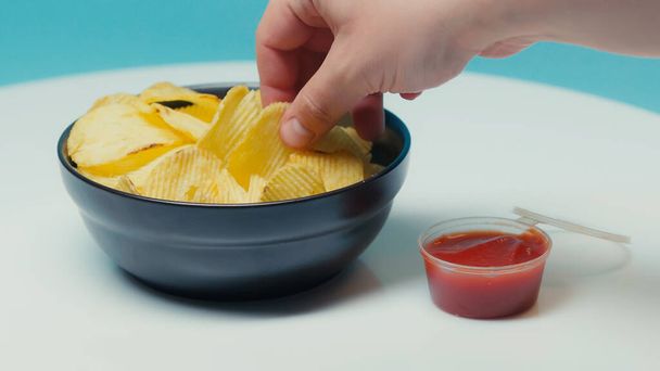 bijgesneden beeld van de mens nemen knapperig geribbelde aardappel chips op blauw - Foto, afbeelding