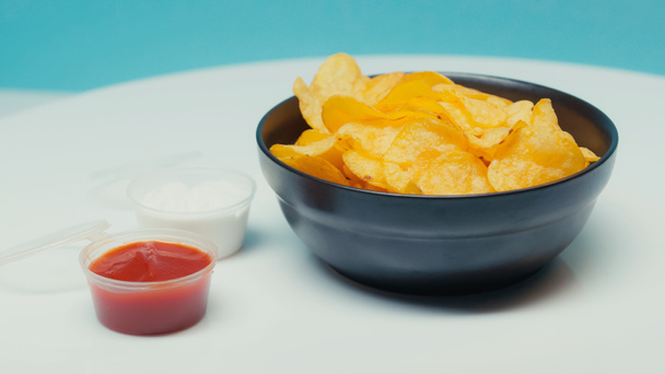 knusprige Kartoffelchips in Schüssel in der Nähe von Plastikbehältern mit Ketchup und Mayonnaise auf blau - Foto, Bild