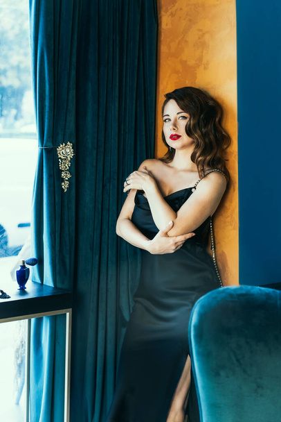Portrait of posh, tempting woman with dark hair in elegant cocktail silk black dress posing in luxury interior. Fashionable and self-confident lady. Celebrity, superstar lifestyle. Vertical card - 写真・画像