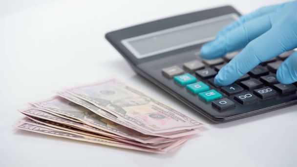 cropped view of person in latex glove using calculator near dollars on white background - Photo, Image