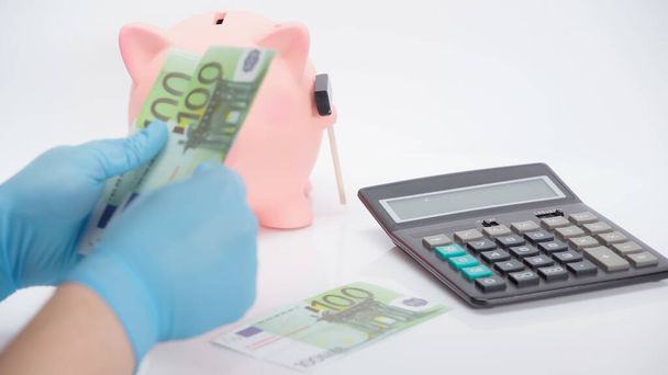 vista recortada de la persona en guantes de látex contando billetes en euros cerca de la calculadora y alcancía sobre fondo blanco  - Foto, imagen