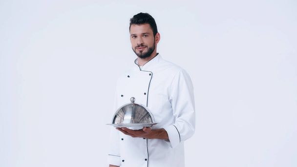 joven chef con plato de servir y cloche mirando a la cámara aislada en blanco - Foto, Imagen