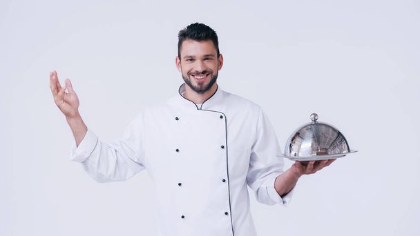 jeune chef avec assiette de service et cloche en acier inoxydable souriant à la caméra isolée sur blanc - Photo, image
