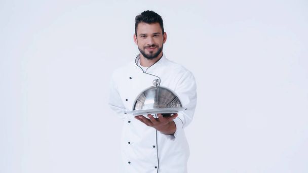 довольный шеф-повар с подачей блюдо и клеш стоя с рукой за спиной изолированы на белом - Фото, изображение