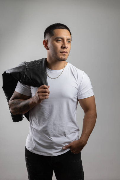 joven modelo masculino vistiendo camiseta blanca, collar y sosteniendo chaqueta de cuero negro, moda urbana e informal, pose de hombre latino, estilo de vida - Foto, Imagen