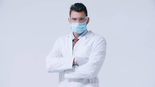 doctor in white coat and medical mask with goggles standing with crossed arms isolated on grey - Fotó, kép