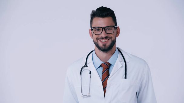 cheerful young doctor in white coat and eyeglasses smiling at camera isolated on grey - Fotó, kép