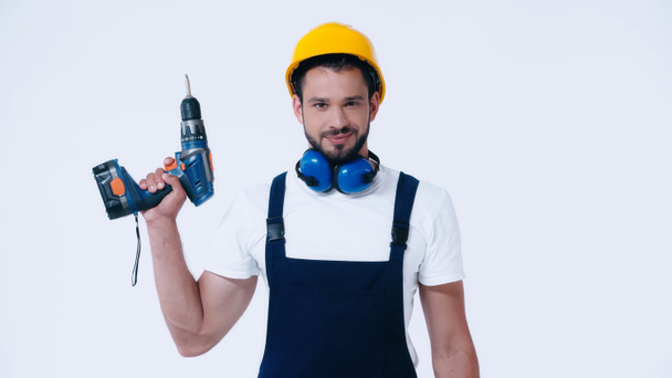 positive builder in hardhat and overalls holding electric drill and looking at camera isolated on white - 写真・画像