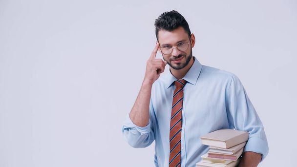 smiling teacher holding pile of books and pointing at head isolated on grey - Photo, Image