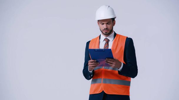 thoughtful engineer in helmet and safety vest looking at clipboard isolated on grey - Foto, afbeelding