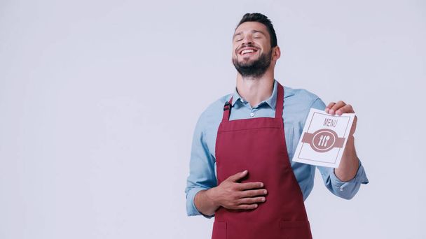 excited waiter laughing and touching tummy while holding menu isolated on grey - Photo, Image