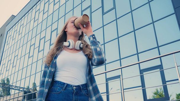 Low angle view of young woman with headphones drinking coffee to go on urban street  - Foto, Bild