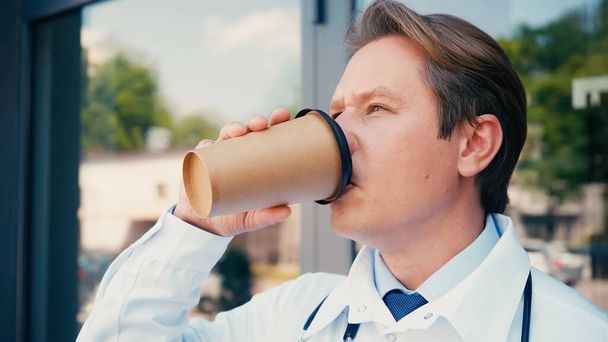 arts in witte jas drinken koffie uit papieren beker buiten - Foto, afbeelding