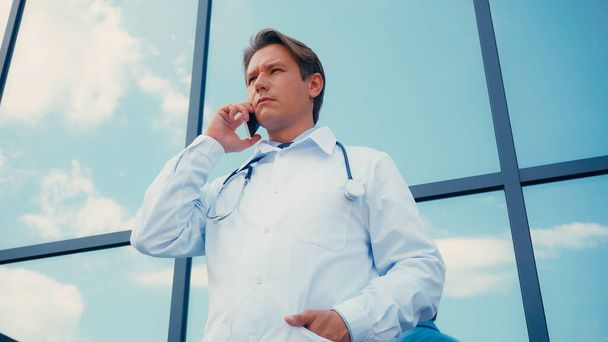 Niedrigwinkel-Ansicht des Arztes mit der Hand in der Tasche des weißen Kittels, der im Freien mit dem Handy spricht - Foto, Bild