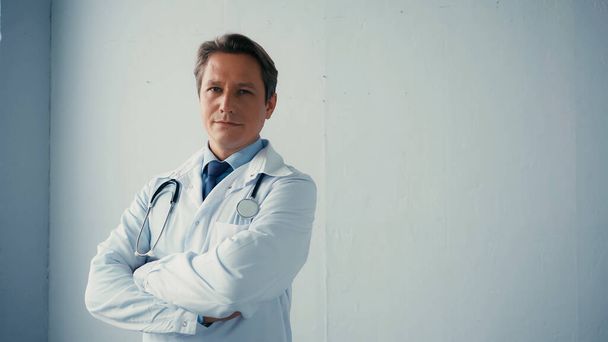  Beyaz önlüklü kendine güvenen doktor gri duvarın yanında çapraz kollarıyla duruyor.  - Fotoğraf, Görsel