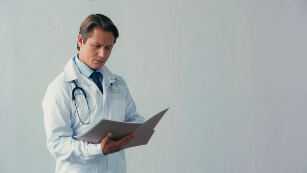 doordachte arts in witte jas op zoek in map met documenten op grijs - Foto, afbeelding