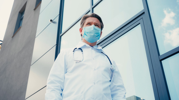 vista de ángulo bajo del médico en bata blanca y máscara médica cerca de la construcción de la clínica - Foto, Imagen