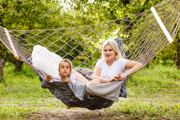 Wide view of a young mother and daughter relaxing together and smiling sitting in a hammock, hugging and lounging during a sunny summer day in a holiday home garden with grass and trees, lifestyle. - Photo, image
