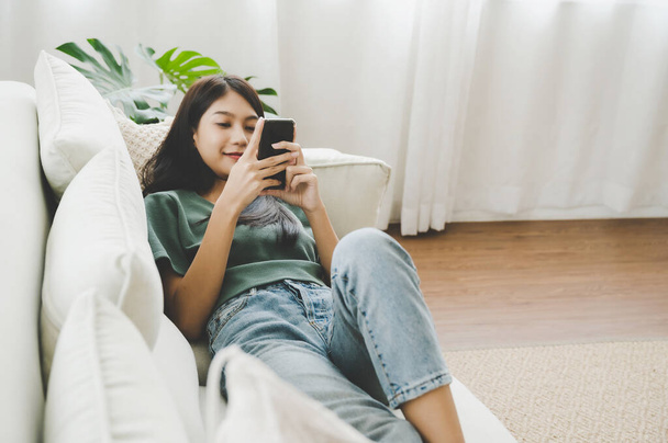 Felice giovane donna asiatica rilassante a casa. Femmina è sdraiato sul divano e utilizzando smartphone mobile - Foto, immagini