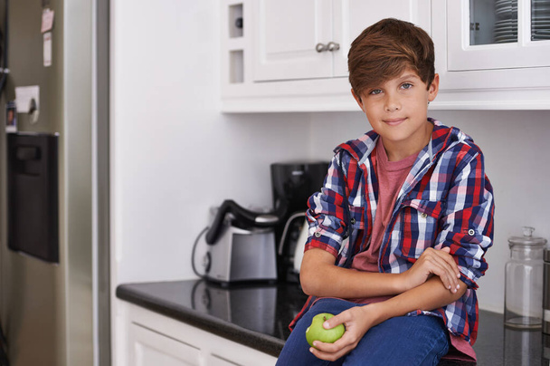 Τα μήλα είναι γιαμ. ένα νεαρό αγόρι κάθεται στον πάγκο της κουζίνας με ένα μήλο στο χέρι του - Φωτογραφία, εικόνα
