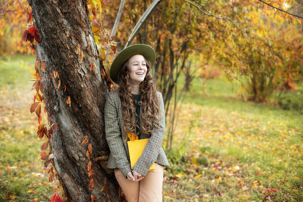Glückliches Mädchen mit grünem Hut und kariertem Sakko, das auf herbstlichem Hintergrund ein Buch liest. Ein lächelndes Mädchen ruht sich im Park aus. Junges Mädchen in Hut und Herbstkleidung steht mit goldenem Herbstlaub gegen Baum. - Foto, Bild