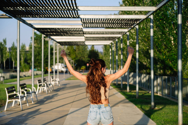 Achteraanzicht van onherkenbare vrouw in gewas top en korte broek staan met opgeheven armen in stadspark en genieten van vrijheid tijdens het zomerweekend - Foto, afbeelding