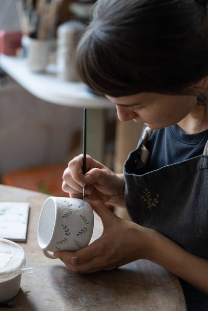 Hands of professional artisan hold white ceramic mug drawing pattern with dark paint using brush on blurred background. Woman enjoys working with handmade pottery craft in studio extreme closeup - Photo, Image