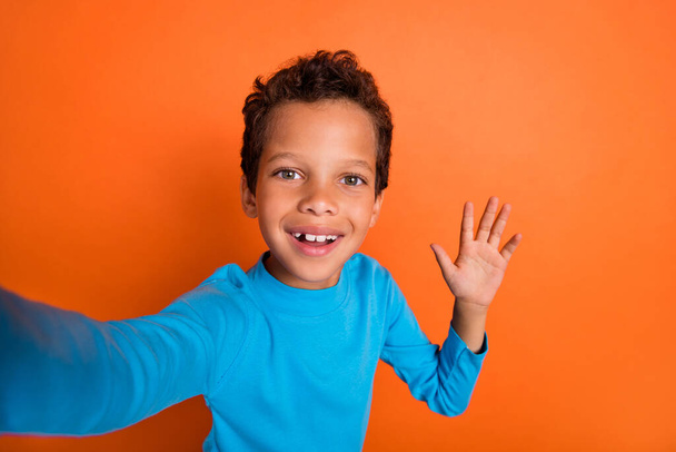 Photo portrait of cute small pupil boy front camera selfie wave hand hello dressed stylish blue garment isolated on orange color background. - Φωτογραφία, εικόνα