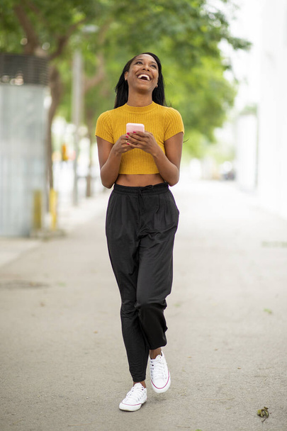 Stylish african american woman laughing while using mobile phone on city street - Foto, Imagem