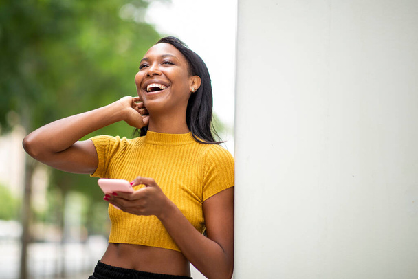 Mulher negra alegre usando telefone celular enquanto se inclina na parede ao ar livre na cidade - Foto, Imagem
