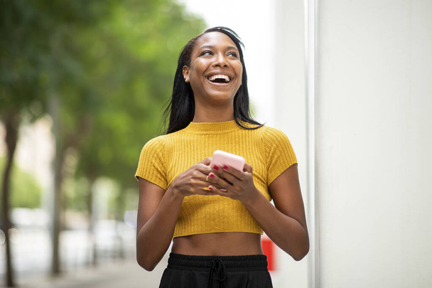 Весела афро-американська жінка, яка озирається і сміється, користуючись мобільним телефоном у місті. - Фото, зображення