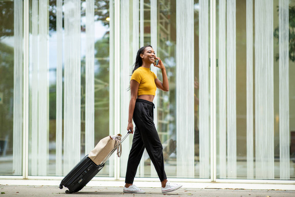 Happy black woman talking on mobile phone while carrying luggage outdoors on city street - Fotoğraf, Görsel