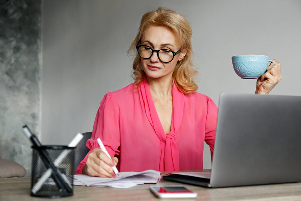 Portrait of adult blonde woman wearing eyeglasses and pink blouse sitting at her workplace with a big blue cup of coffee. Teacher grading papers. Background, copy space, close up. - Foto, Imagem