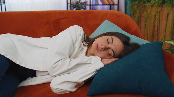 Tired caucasian adult girl lying down in bed taking a rest at home. Carefree young woman napping, falling asleep on comfortable orange sofa with pillows. Closed her eyes enjoy daytime nap alone - Foto, Bild