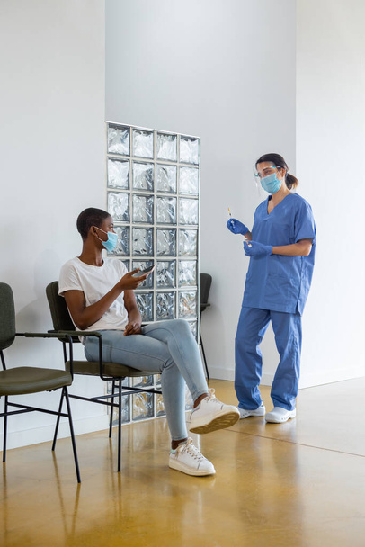 Young African American female patient using on smartphone in a waiting room, talking to female doctor in protective uniform, latex gloves, face mask before vaccinating in clinic, coronavirus outbreak. - Valokuva, kuva