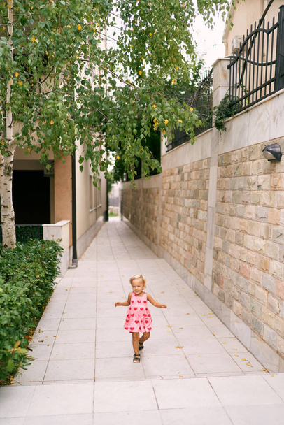 Little girl runs along a paved path in the garden near the house waving her arms. High quality photo - Фото, изображение
