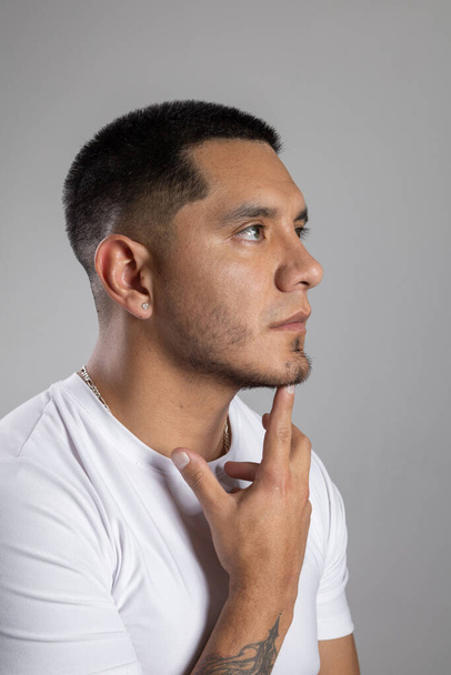 Лицо молодого латинского человека с короткими городскими волосами с бородой и белой футболкой в студии с указательным пальцем на подбородке, думая: - Фото, изображение