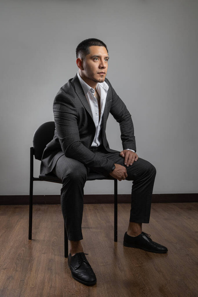 young latin man with short hair and beard, business clothes, suit, shirt and tie, fashion and lifestyle in studio sitting on a chair - Fotografie, Obrázek