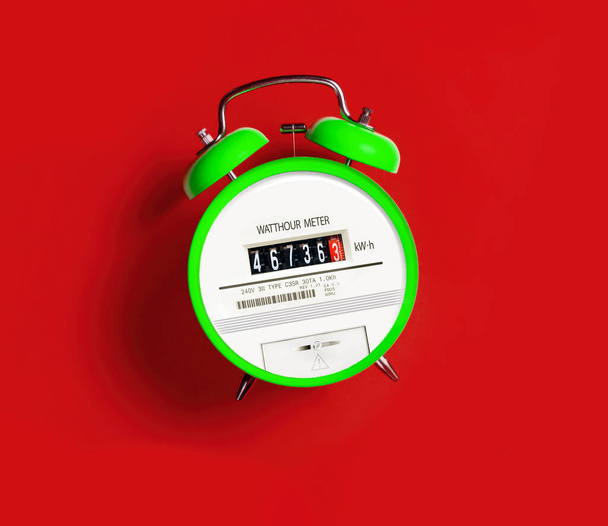 Электросчетчик с будильником на красном фоне. Пора платить за коммунальные услуги. Увеличение расходов на электроэнергию - Фото, изображение