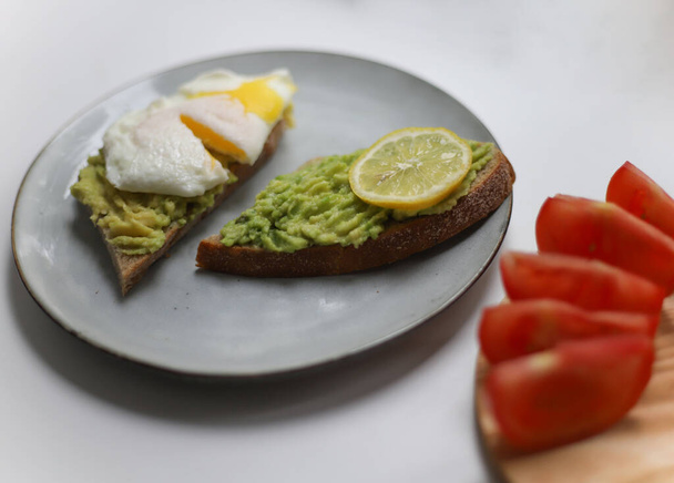 two black bread sandwiches smeared with ripe avocado and topped with scrambled eggs and yellow lemon lie on a gray round kitchen plate next to pieces of a red tomato from the garden. for banners of flyers of cookbooks of recipes and notebooks - Photo, Image