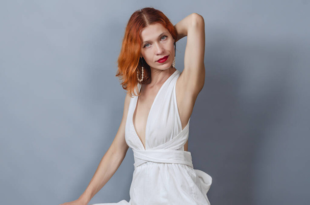 Portrait of red-haired woman in stylish off-shoulder white dress. Looks into camera. Hand is raised behind head. - Photo, Image