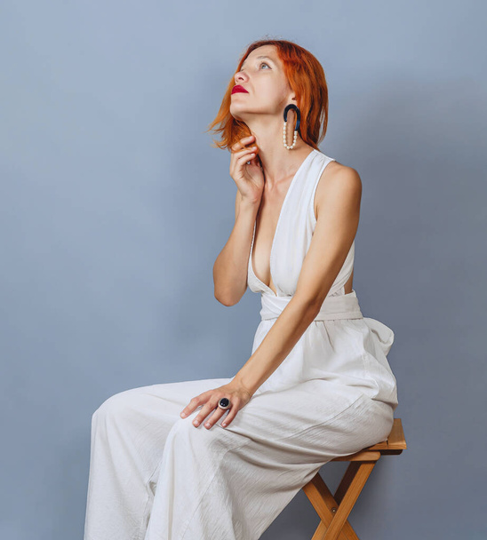 Red-haired woman with dreamy expression, focused to side, thinking about vacation after work, wearing an elegant white dress, has red hair. Posing on gray background. - Photo, Image