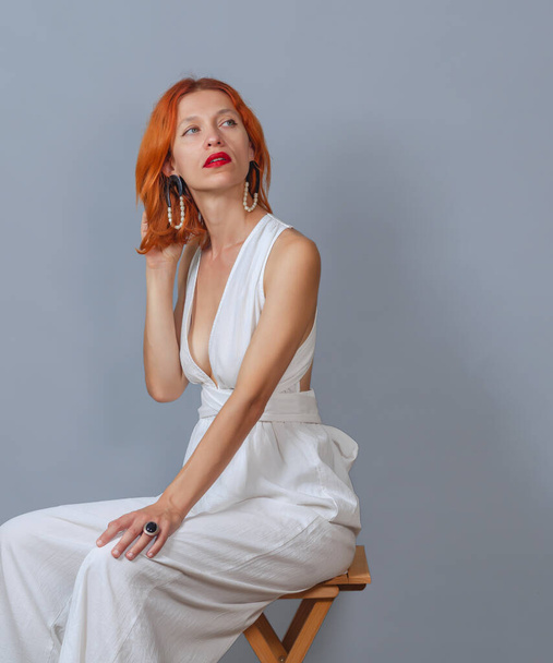 Red-haired woman with dreamy expression, focused to side, wearing elegant white dress, has red hair. Posing on gray background. - Photo, image