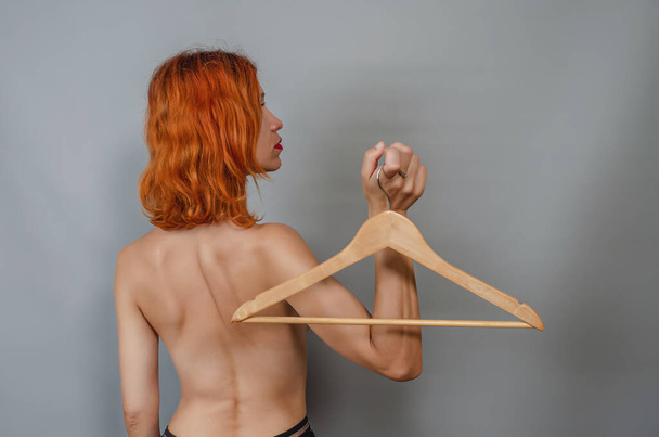 Jonge roodharige naakte vrouw houdt een lege houten hanger in haar hand. Achteraanzicht. Concept van niets te dragen van kleren. Vrouw denkt over het veranderen van kleding in haar garderobe. - Foto, afbeelding