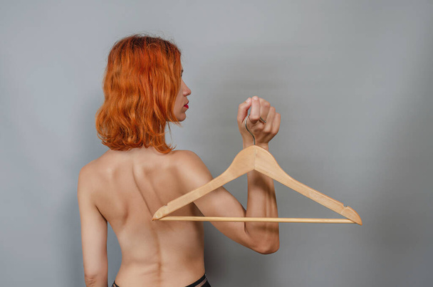 Young red-haired naked woman holds an empty wooden hanger in her hand. Back view. Concept of nothing to wear from clothes. Woman is thinking about changing clothes in her wardrobe. - Zdjęcie, obraz