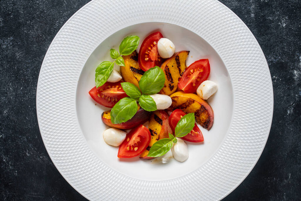 Grilled peach salad with mozzarella, tomato, green basil and sauce. Fresh gourmet salad in white plate on black background, close up, top view - Photo, Image