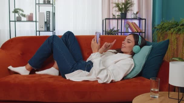 Νεαρή γυναίκα με ασύρματα ακουστικά χαλαρώνοντας ξαπλωμένη στον καναπέ στο σπίτι επιλέγοντας ακρόαση αγαπημένο ενεργητική disco rock n roll χορευτική μουσική στο smartphone. Δραστηριότητες Σαββατοκύριακου. Χαρούμενο χαρούμενο κορίτσι - Πλάνα, βίντεο