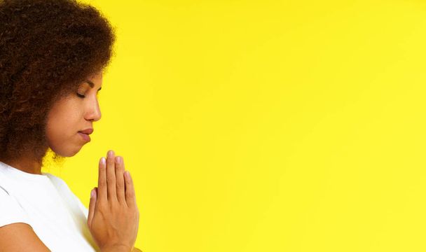 黄色の背景に隔離された白いTシャツを着て祈りの位置に若いアフリカ系アメリカ人女性。アフリカの少女は神のスタジオショットを祈る。精神的な女性が祈りに手を置く.  - 写真・画像