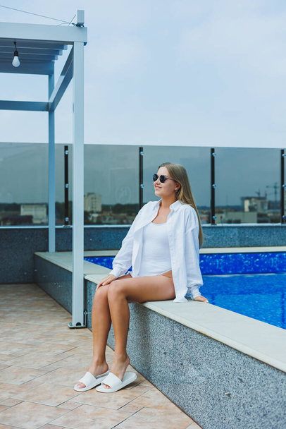 A cheerful young woman in a white swimsuit and a white shirt is sitting by the pool. A woman in sunglasses and a swimsuit. Summer vacation by the pool. Tanned girl - 写真・画像