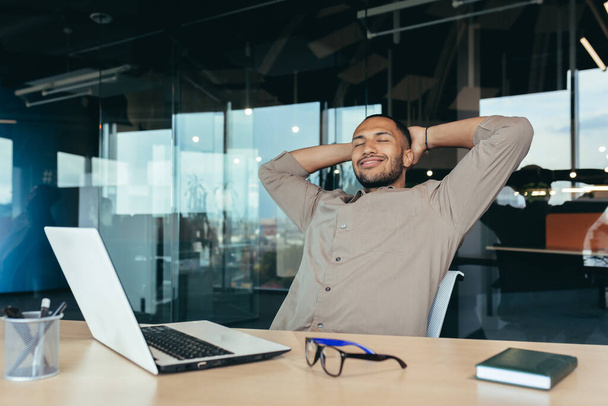 mixed race man relaxing in office with hands behind head looking out window and smiling, businessman daydreaming inside modern office building, successful investor happy with work. - Foto, Imagem